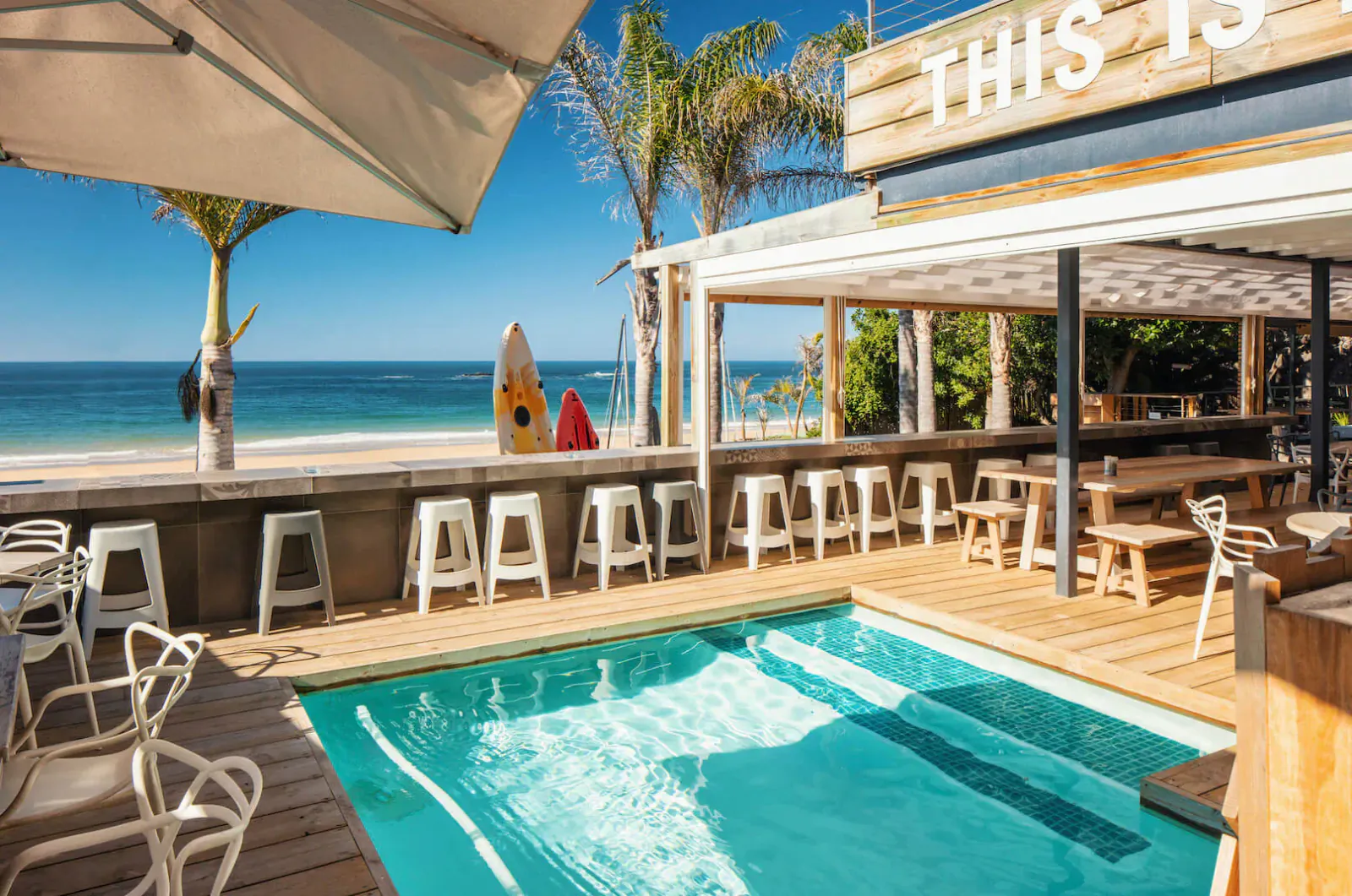 The Bungalow seaside venue in Plettenberg Bay with infinity pool, beach view, surfboards, and 'THIS IS' sign.