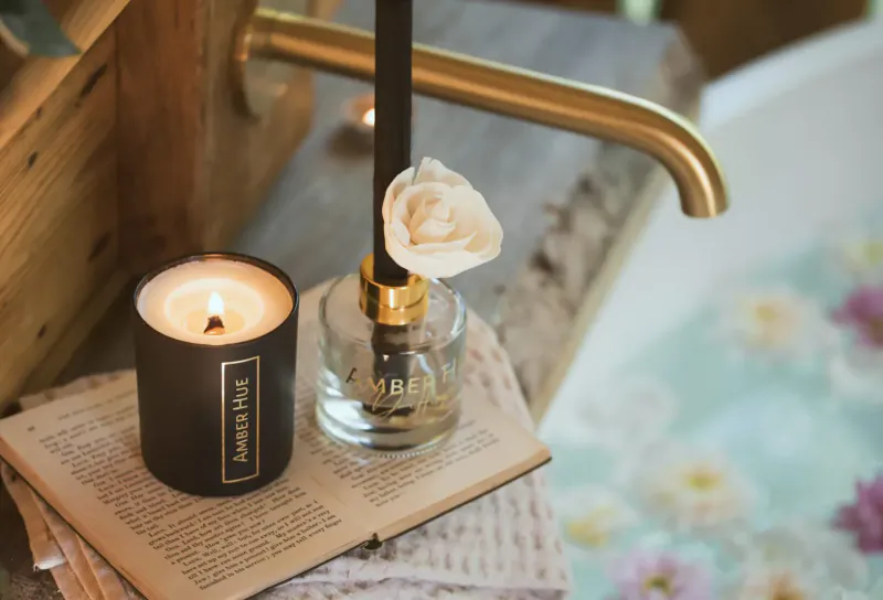Luxury bathroom setup with lit Amber Hue candle, reed diffuser with white rose, brass faucet, book, and pink bath flowers