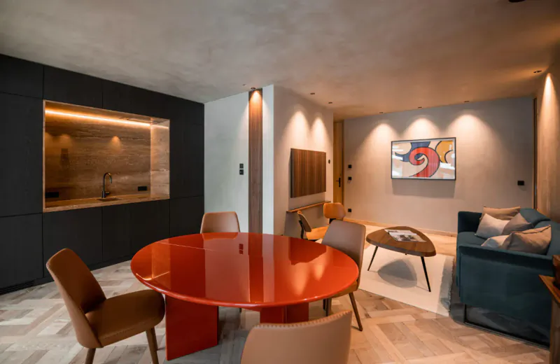 Modern luxury chalet living room with red round dining table, teal sofa, abstract wall art, and warm lighting.