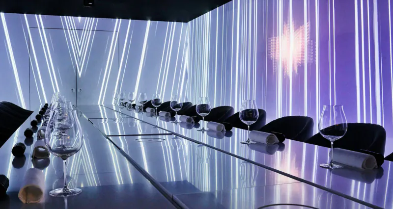 Elegant long dining table set in Monarch Theatre with wine glasses, napkins, and glowing white-purple LED curtains.
