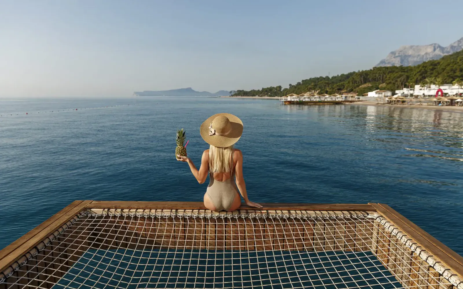 Woman in white bikini and straw hat holding green bottle, sitting on wooden net platform overlooking sea and mountains
