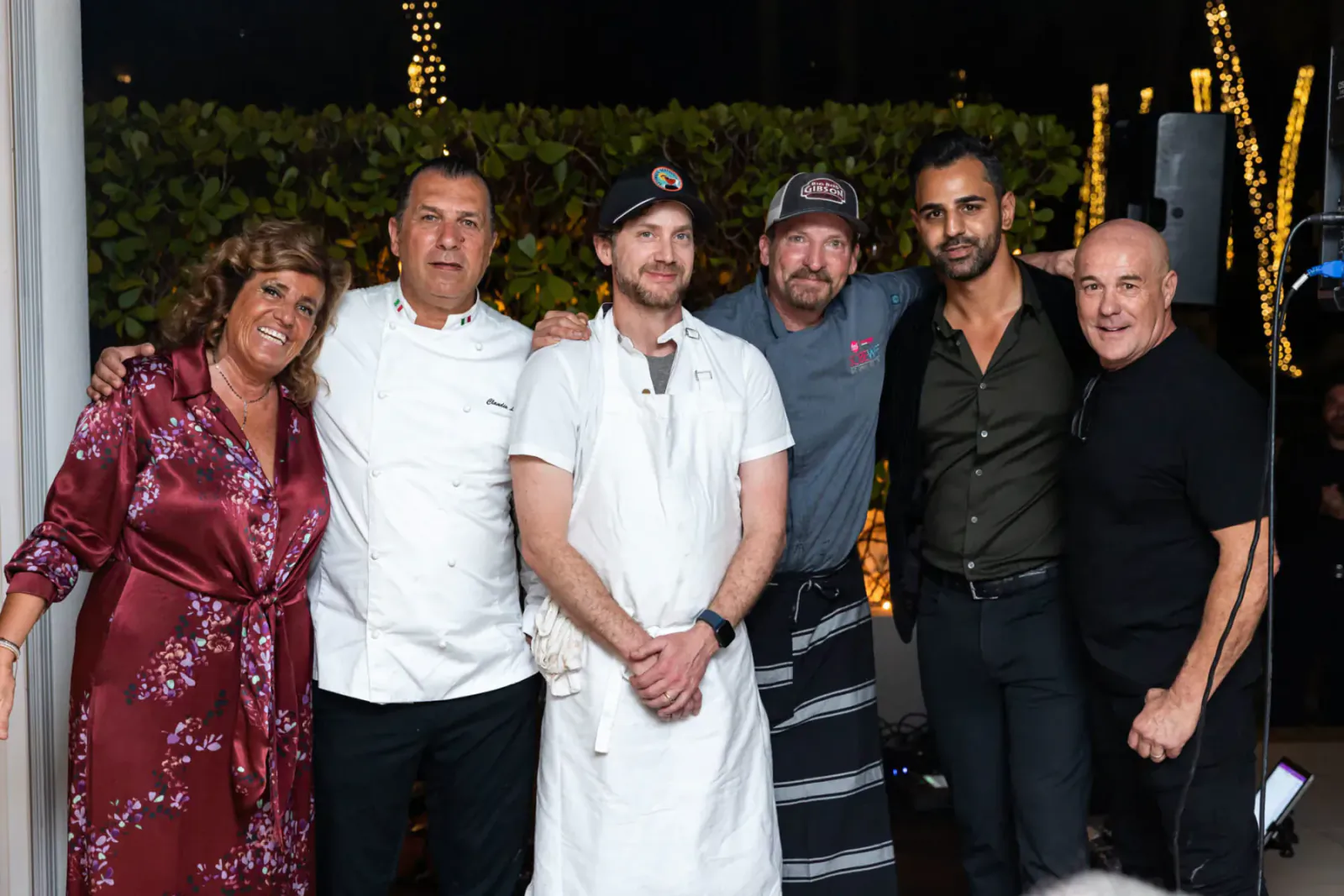 Group of five smiling at BBQ Marvel event on festoon-lit patio: woman in red robe, three chefs in whites, man in black shirt.