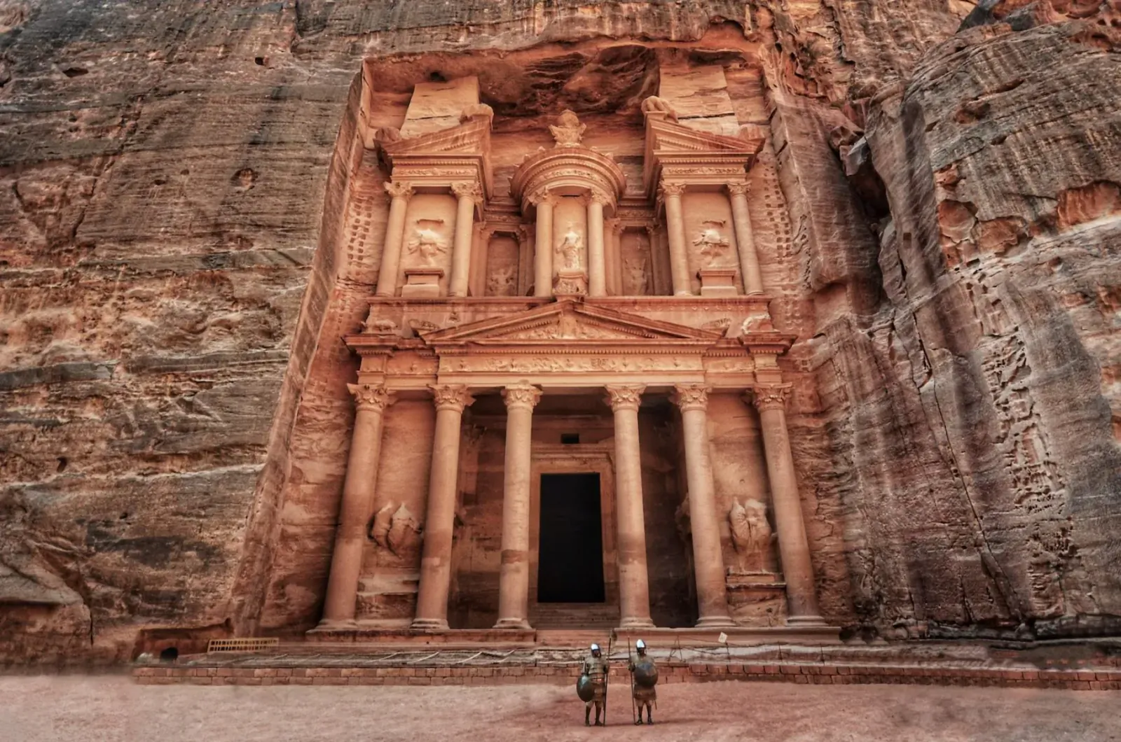 Two hikers stand before the grand Treasury (Al-Khazneh) carved into Petra's rose-red cliffs, Jordan.