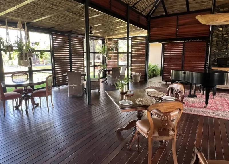 Spacious luxury African lodge interior with wooden floors, antique chairs, tables set for dining, grand piano, and lush plants.