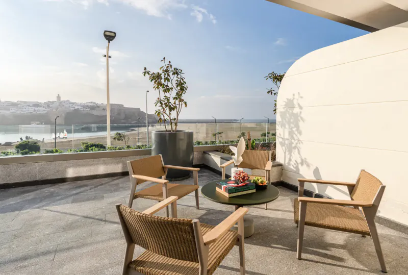 Fairmont La Marina Rabat-Salé Hotel rooftop terrace with wicker chairs around low table with flowers, potted plants, overlooking marina and cityscape.