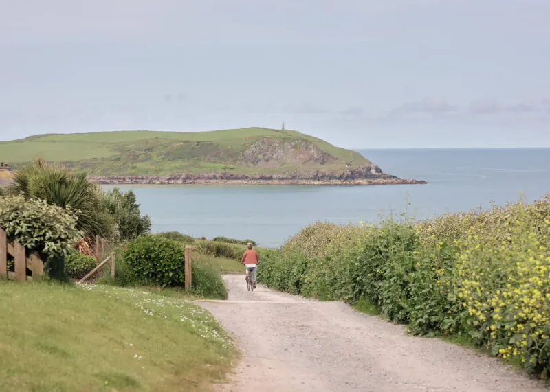 Person cycling on gravel path through green hedges toward coastal headland with lighthouse at St Moritz Hotel, Cornwall