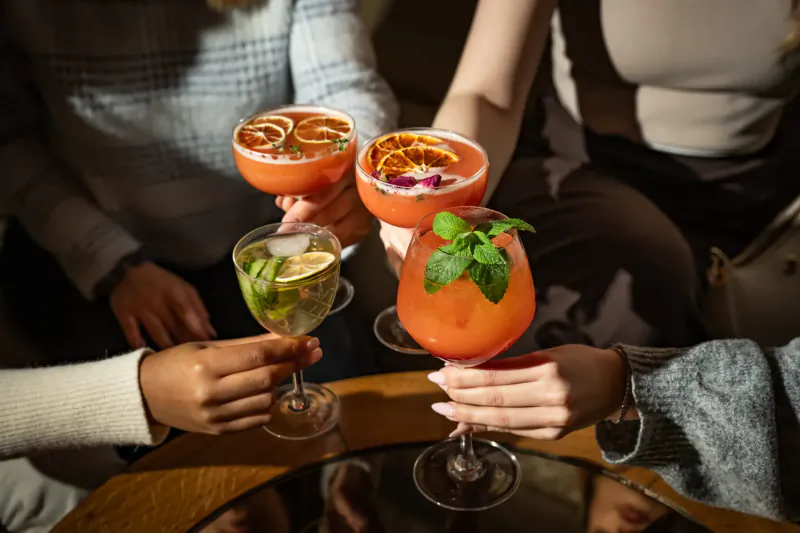 Four women clinking colorful cocktails garnished with orange slices, mint, and rose petals over a table at Mint Leaf London gallery.