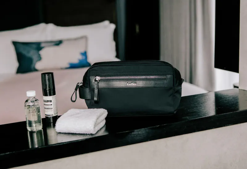 Black Antler Fenchurch dopp kit on hotel bedside table with toiletries, white towel, and pillows in background