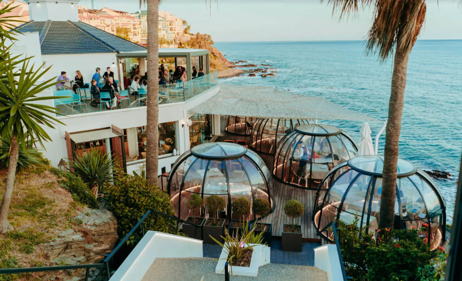 Yucas Mare & Mar de Copas seaside venue with glass dome pods on cliff overlooking ocean, people lounging on balcony amid palms.