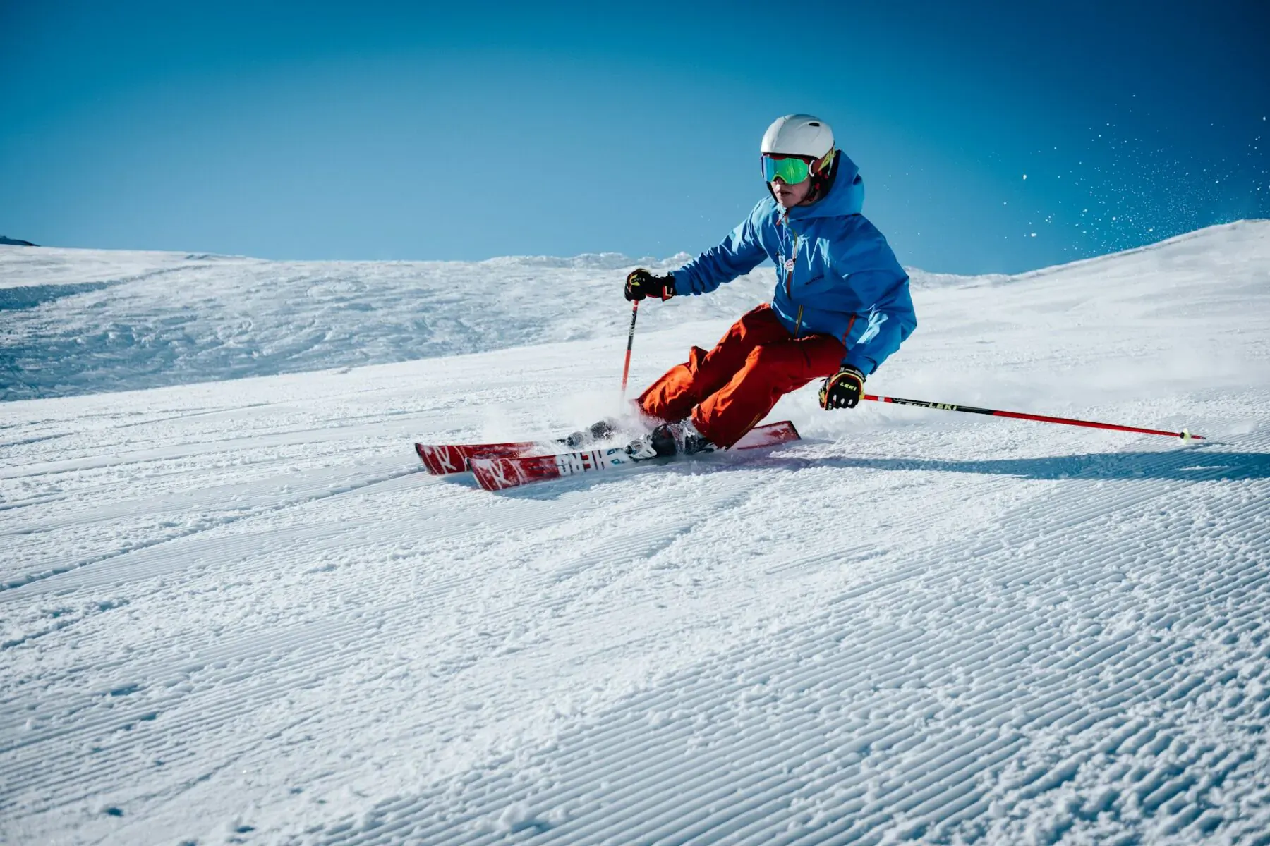 Advanced skier in blue jacket and red pants carves down snowy mountain slope under clear blue sky.