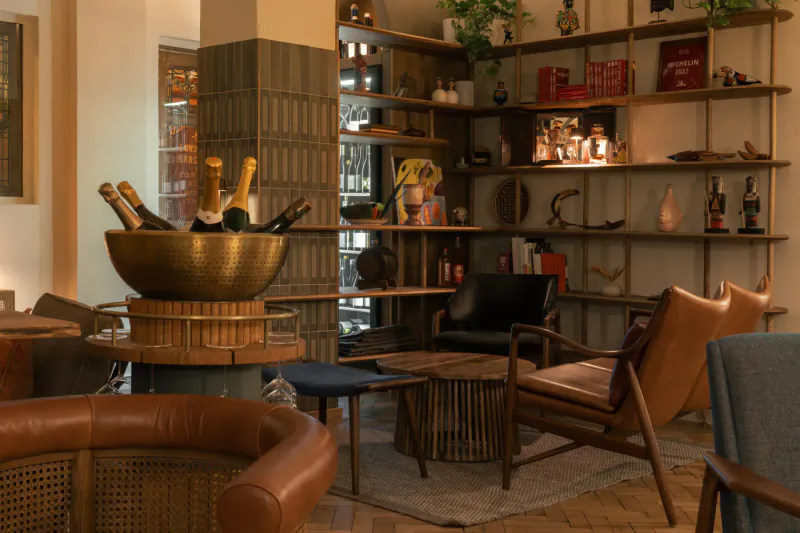 Elegant lounge interior with gold ice bucket of champagne bottles, wooden shelves of liquor and books, leather chairs, plants.