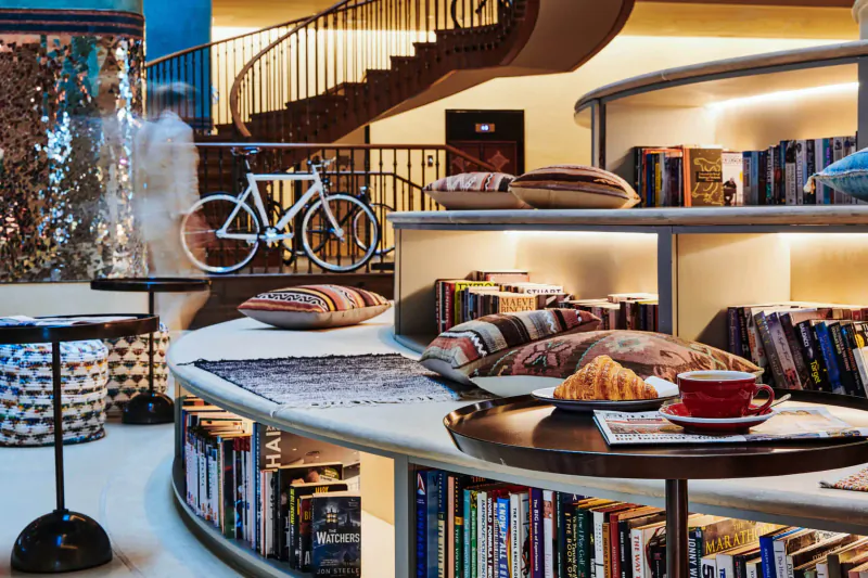 Cozy 25hours Hotel Nomad Hotspot in Dubai: curved bookshelves with pillows, white bicycle, croissant and coffee on table by spiral stairs.