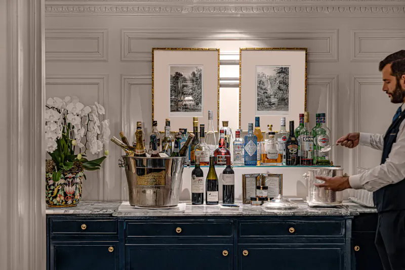 Bartender in vest pouring drink from ice bucket at elegant bar with liquor bottles, white orchids, and framed art in luxurious room