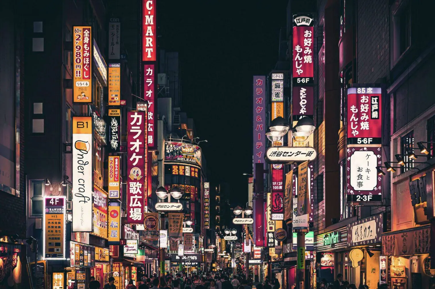 Vibrant neon-lit Tokyo street at night with Japanese signs like C-mart and colorful billboards.