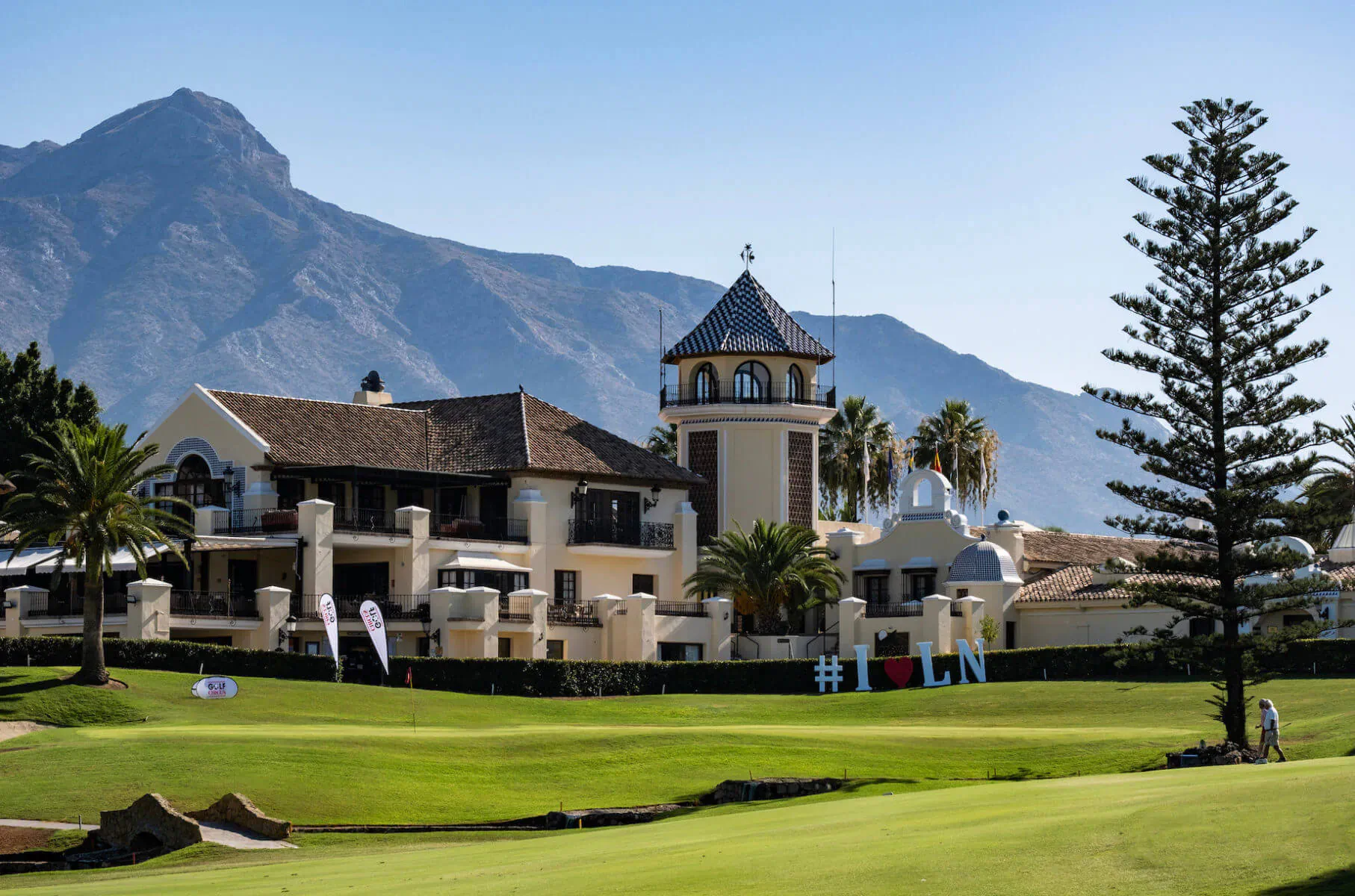 Los Naranjos Golf Club: luxurious clubhouse with tower, palm trees, and golf course against Marbella mountains.