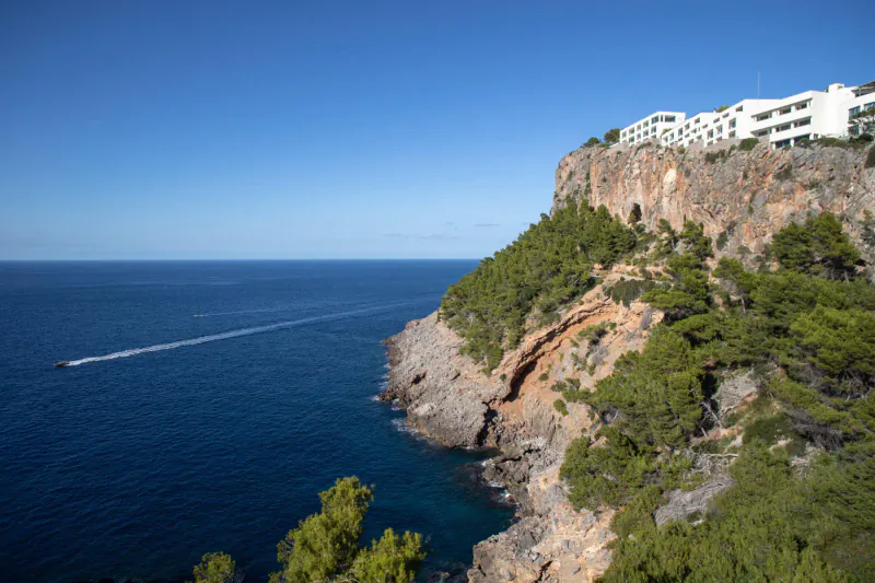 White Jumeirah Mallorca hotel atop rocky cliff overlooking sea with speedboat trail, pines and blue sky.