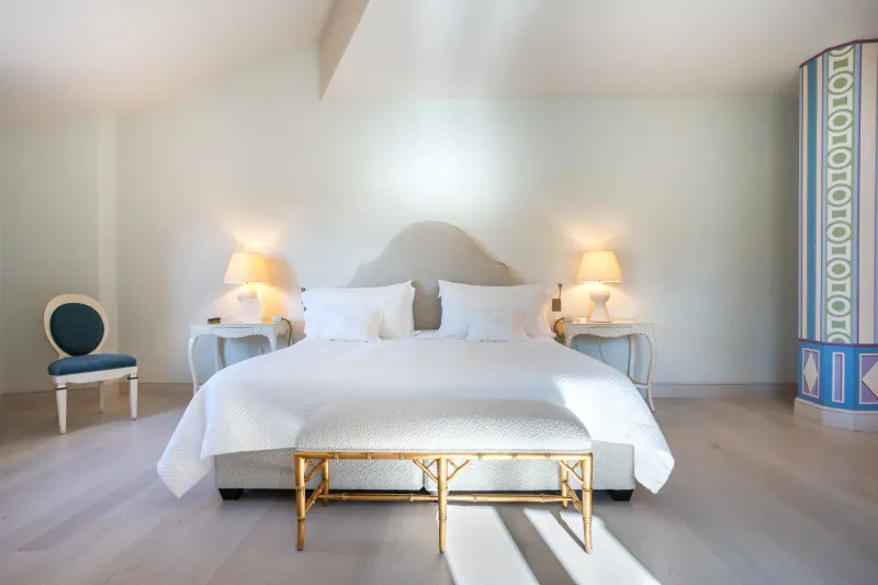 Elegant hotel bedroom with white king bed, gold bench, curved green chair, lamps, and blue patterned pillar.