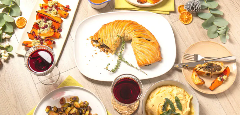 Christmas dinner spread: sliced orange pastry with herbs, roasted veg, mash, sprouts, carrots, red wine on wooden table with eucalyptus.