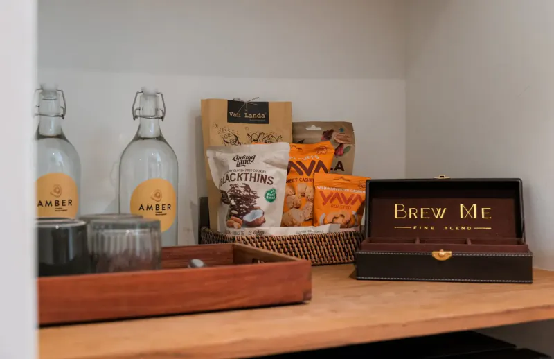 Hotel shelf with Amber water bottles, snack bags like The Larder and Svasa, Brew Me tea box, and glass in tray.