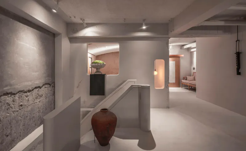 Stylish contemporary restaurant interior with white walls, exposed concrete, potted plant, large terracotta vase, staircase, and lounge seating.