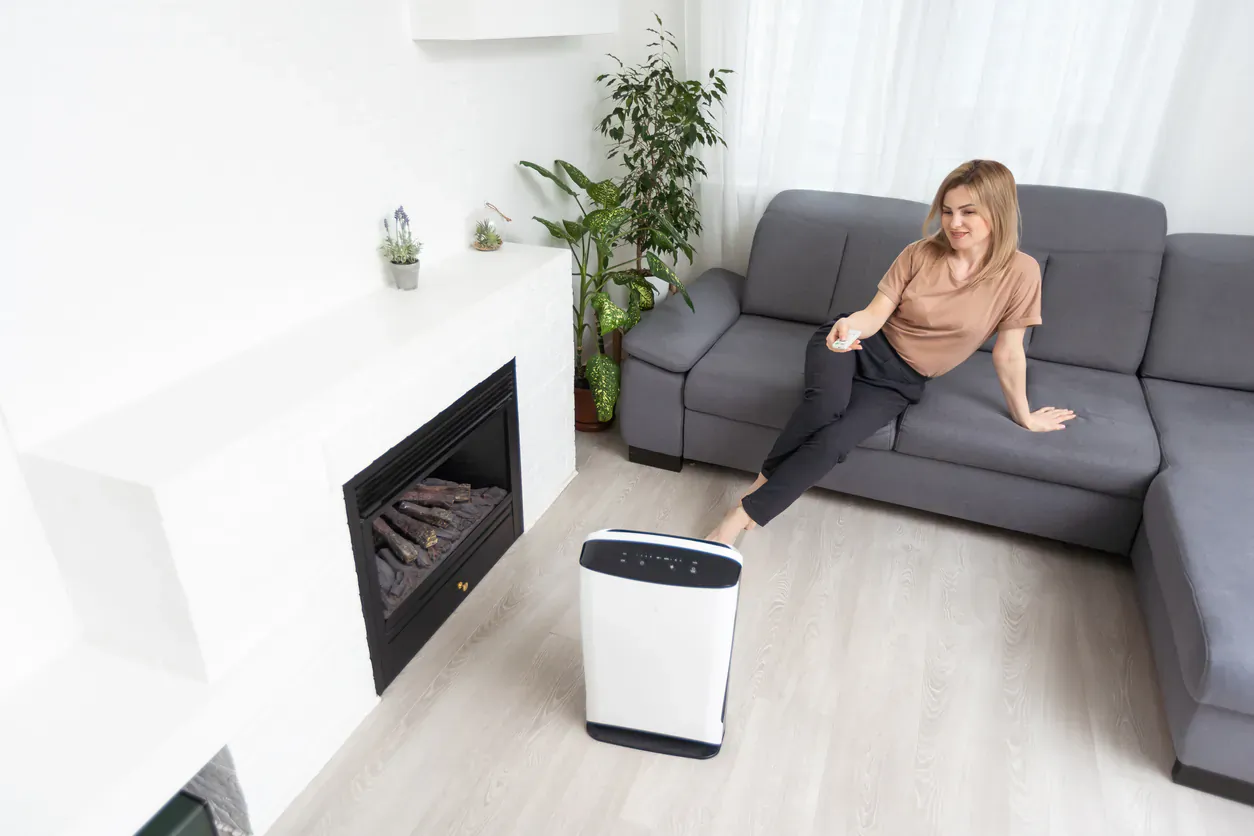 Woman relaxing on gray sofa with legs propped on white air purifier in modern living room by fireplace.