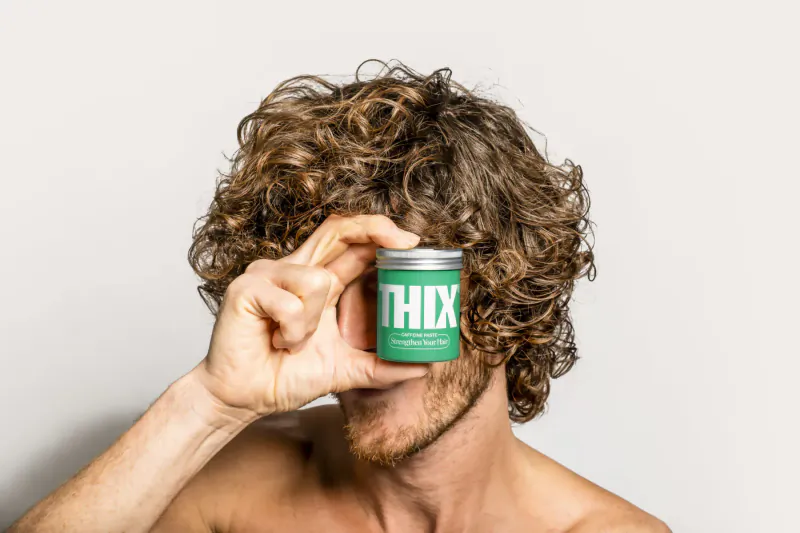 Man with curly hair holds small green THIX jar over one eye, shirtless on white background