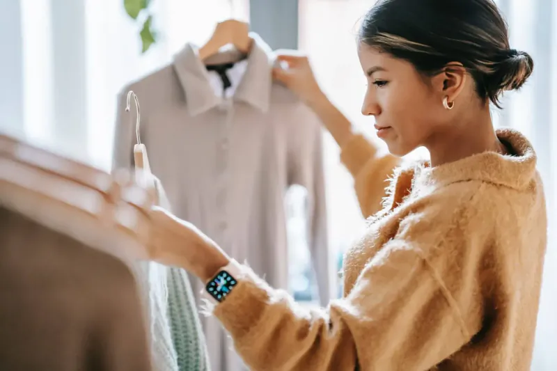 Asian woman in orange cardigan examines light gray shirt on hanger in boutique, spring fashion