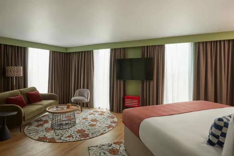 Cozy Hotel Indigo room with green sofa, round rug, TV, and king bed by large curtained windows.