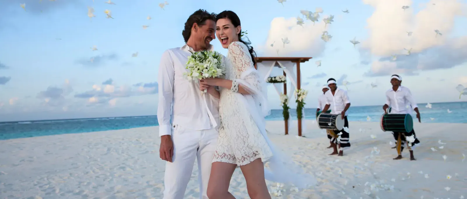 Bride in lace gown and groom in white suit embrace under floral arch on Maldives beach, with musicians and petals.