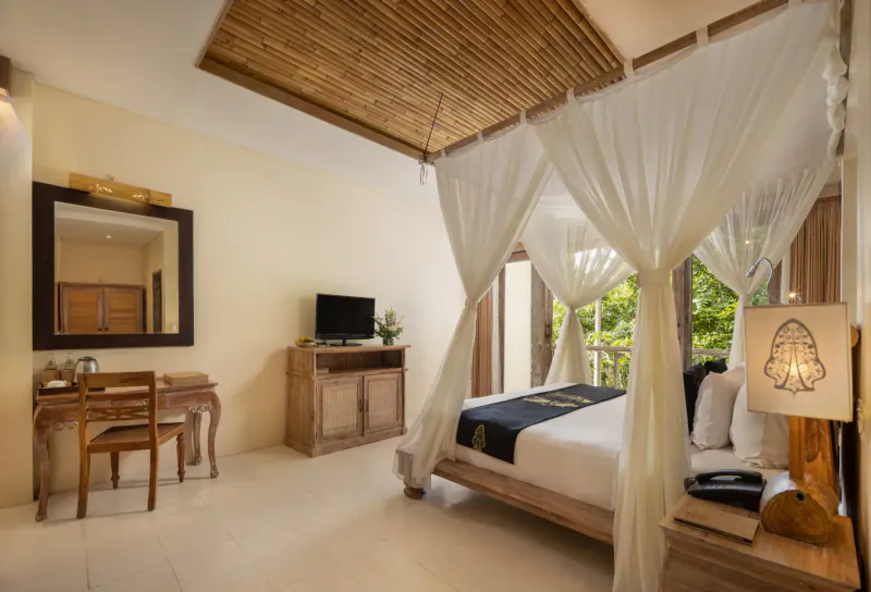 Luxurious Balinese hotel room with white canopy bed, bamboo ceiling, wooden desk, TV, and garden view.