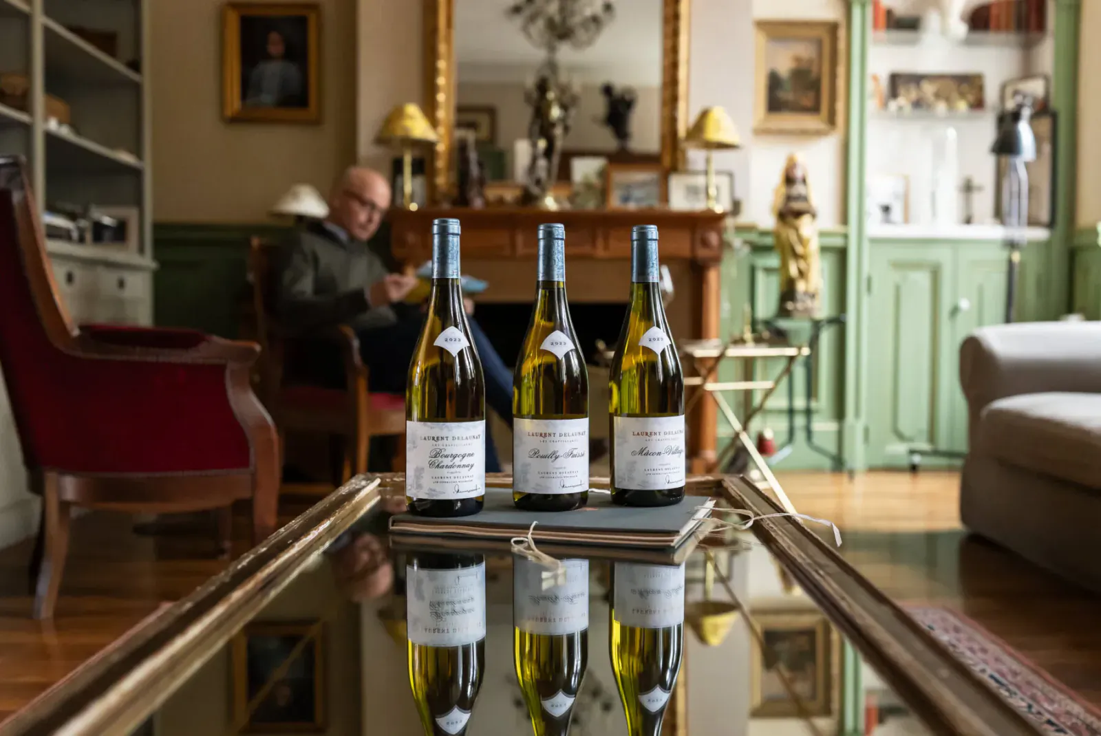 Three French white wine bottles on a table in elegant room, man seated nearby, Christmas decor, festive setting.