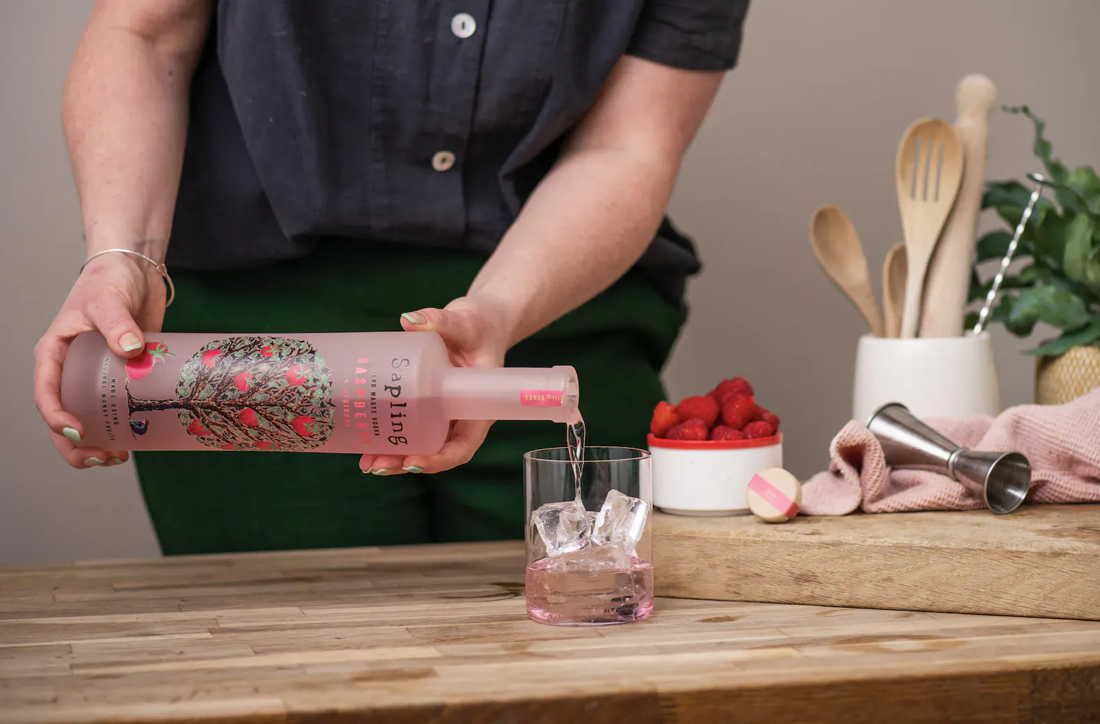 Woman pouring pink Sapling Raspberry & Hibiscus Vodka from bottle into ice-filled glass with strawberries on wooden counter.