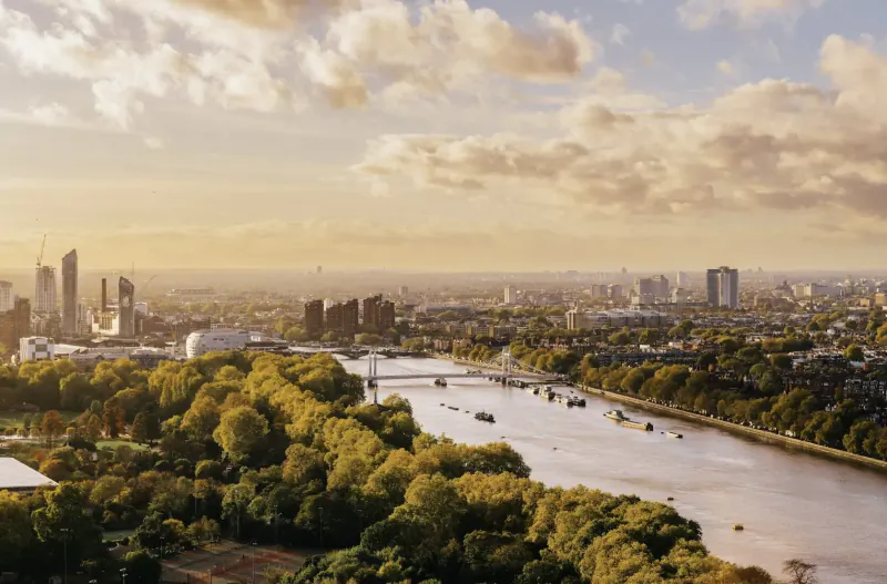 Golden hour aerial panorama of Battersea Power Station, River Thames, and London skyline with trees and boats.