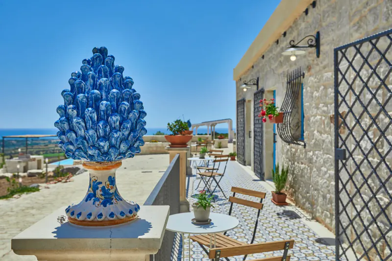 Blue ceramic pine cone sculpture on stone terrace of luxury Sicilian villa with sea view, chairs, plants, and pool.