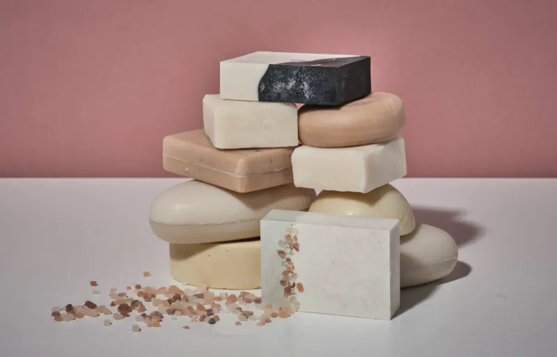 Stack of assorted natural soap bars in white, beige, black, and pink hues with scattered bath salts on white surface