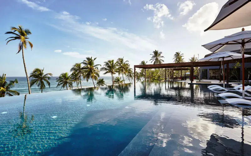 Infinity pool at Ani Private Resorts overlooking tropical beach with palm trees, umbrellas, and sun loungers under blue sky.