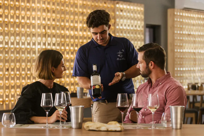 Server pouring white wine for smiling couple at wooden table with glasses and bread, golden wine wall backdrop