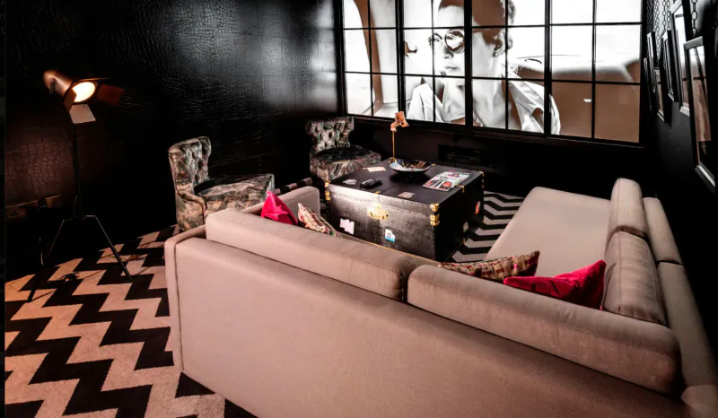 Plush club bar lounge in Hotel Gotham with black zig-zag floor, beige sofa, armchairs, vintage trunk table, and wall mural of woman.