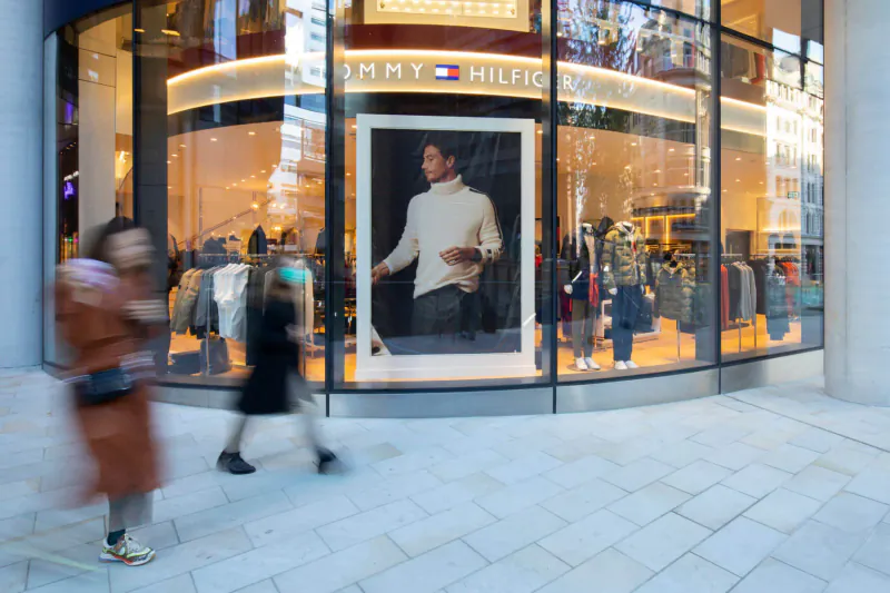 Pedestrians walk past Tommy Hilfiger store window with large ad of man in cream sweater amid clothing displays.