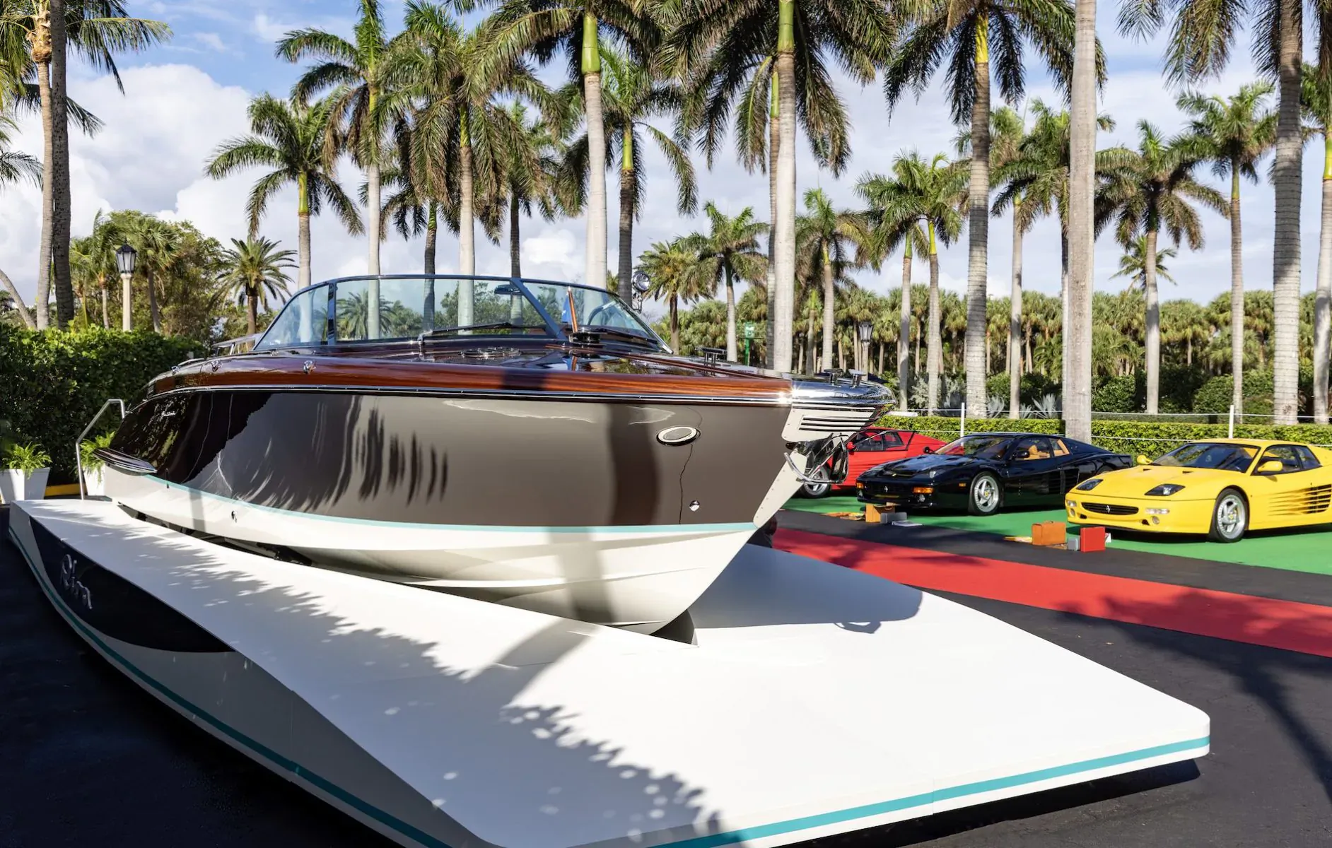 Brown Riva speedboat on platform with red Ferrari and yellow Lamborghini at Palm Beach Cavallino Classic amid palm trees.