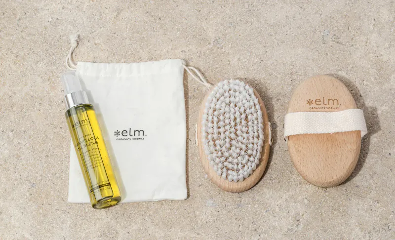 Elm Organics dry brush with white strap, white pouch, and yellow oil spray bottle on beige surface