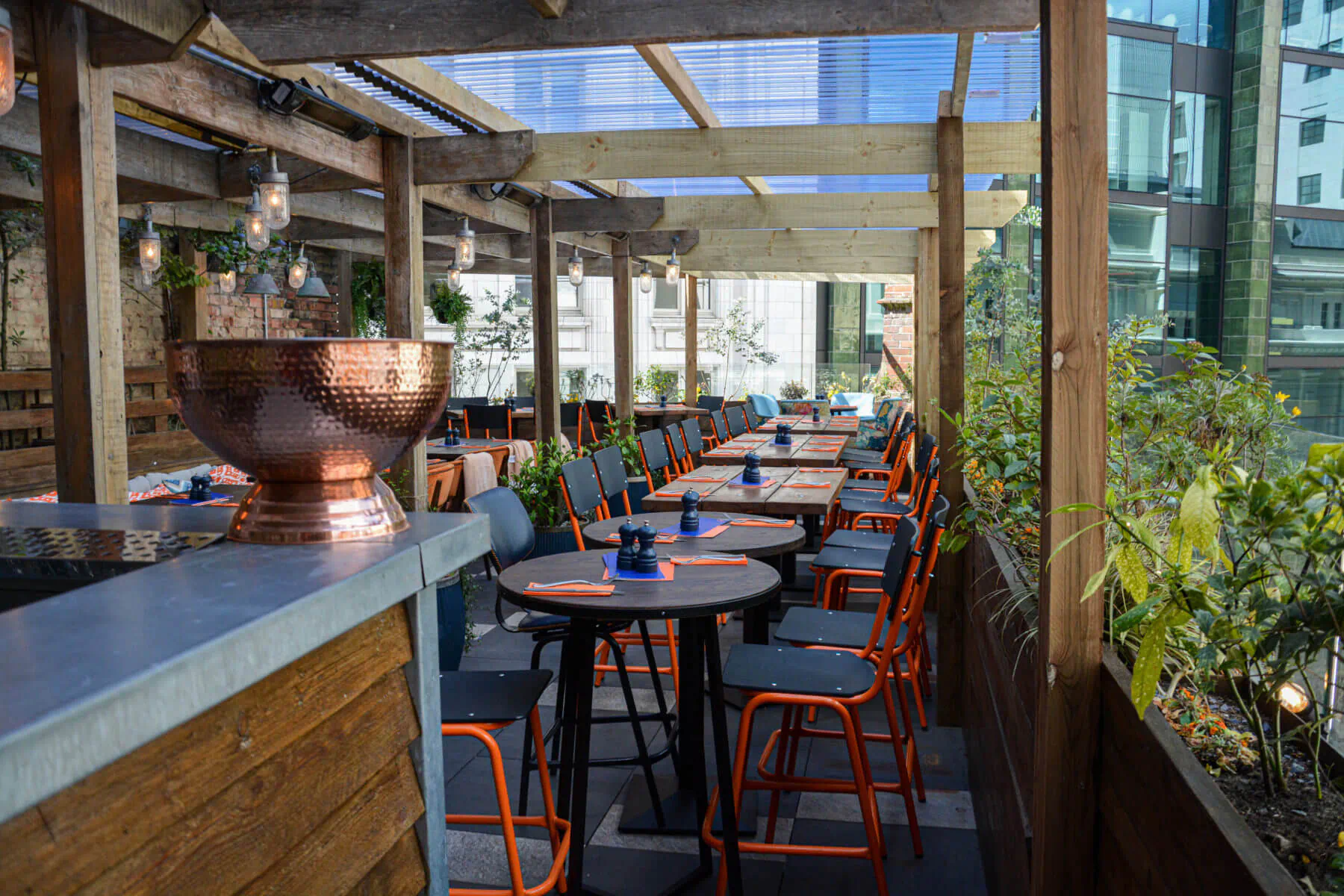 CoqBull Soho rooftop terrace restaurant with copper bowl on bar, orange chairs, round tables, plants, city view.