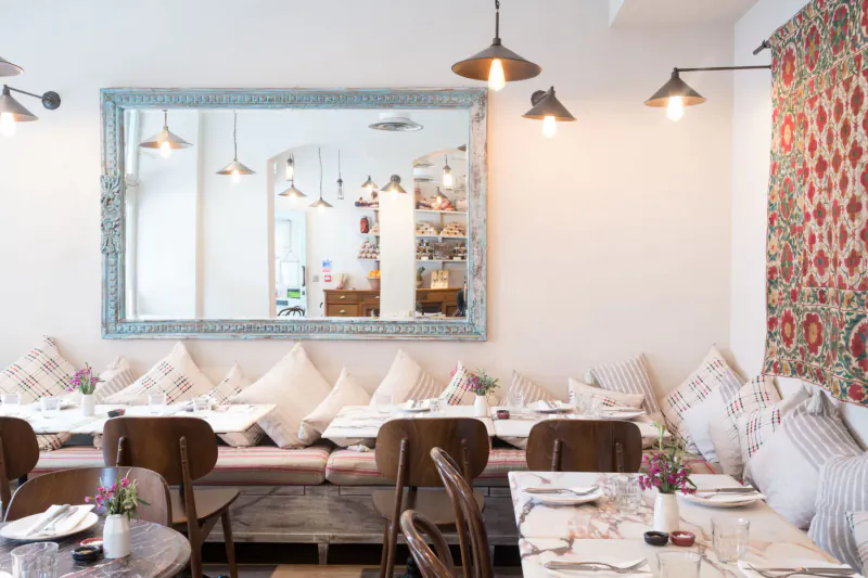 Cozy Villa Mamas restaurant interior in London with cushioned bench seating, wooden chairs, white tables, pendant lights, large mirror, and floral wall tapestry.