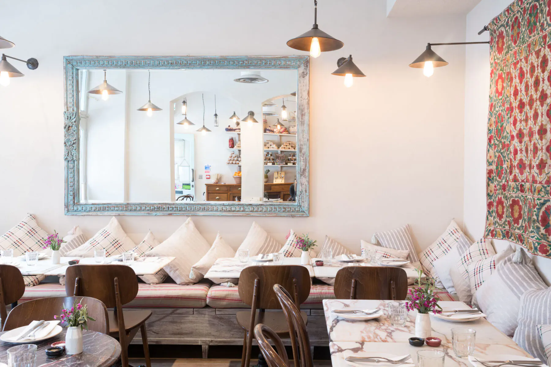 Cozy Villa Mamas restaurant interior in London with cushioned bench seating, wooden chairs, white tables, pendant lights, large mirror, and floral wall tapestry.