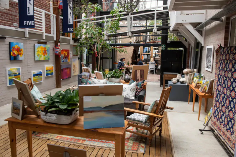 Indoor gallery at The Lofts Boutique Hotel with colorful paintings, plants, wooden furniture, and people in atrium courtyard.
