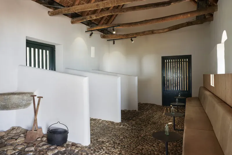 Interior of Clara’s Barn at Vergenoegd Löw: white walls, wooden beams, green doors, stone bench, basin, and cauldron on pebble floor.