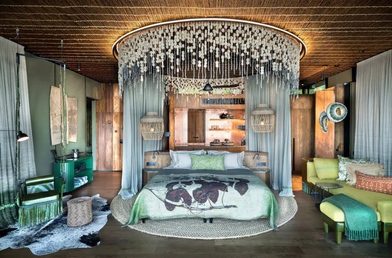 Luxurious Lolebezi safari lodge bedroom with leaf-patterned bed, shell chandelier, green curtains, and eclectic decor.