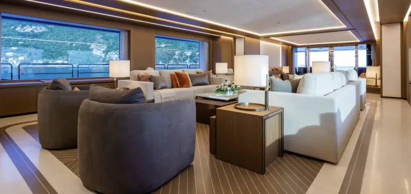 Luxurious yacht lounge with white sofas, lamps, and large windows overlooking lake and mountains.