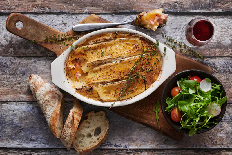 Baked eggplant gratin topped with cheese and thyme on wooden board with baguette, salad, and red wine glass