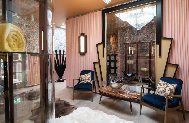 Art deco hotel lounge with pink walls, large mirror, blue armchairs, wooden table, and potted plants